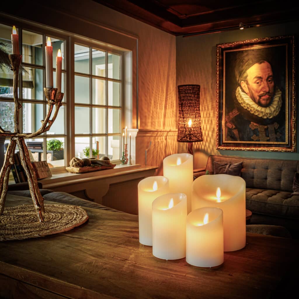 Een gezellige woonkamer met een LED wax kaars D12,5 X H15 CM oplaadbaar warm wit met afstandsbediening gloeiend op een houten tafel, een kandelaar met hoge kaarsen, een groot historisch portret aan de muur en een bank bij het zonverlichte raam.