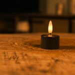 Eine kleine, schwarze, flammenlose LED-Kerze steht auf einem rustikalen Holztisch und leuchtet sanft in einem warm beleuchteten Raum mit verschwommenen Hintergrundmöbeln.