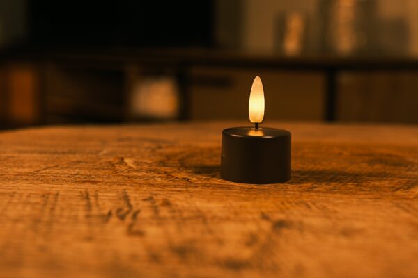 Eine kleine, schwarze, flammenlose LED-Kerze steht auf einem rustikalen Holztisch und leuchtet sanft in einem warm beleuchteten Raum mit verschwommenen Hintergrundmöbeln.