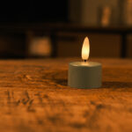 Eine kleine, batteriebetriebene Teelichtkerze mit einer warmen, gelben Flamme steht auf einem Holztisch. Der Hintergrund ist sanft verschwommen und schafft eine gemütliche und ruhige Atmosphäre.