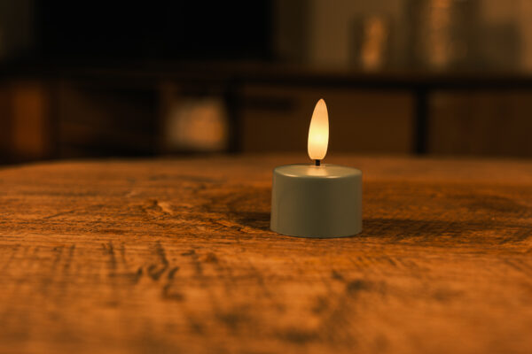 Eine kleine, batteriebetriebene Teelichtkerze mit einer warmen, gelben Flamme steht auf einem Holztisch. Der Hintergrund ist sanft verschwommen und schafft eine gemütliche und ruhige Atmosphäre.